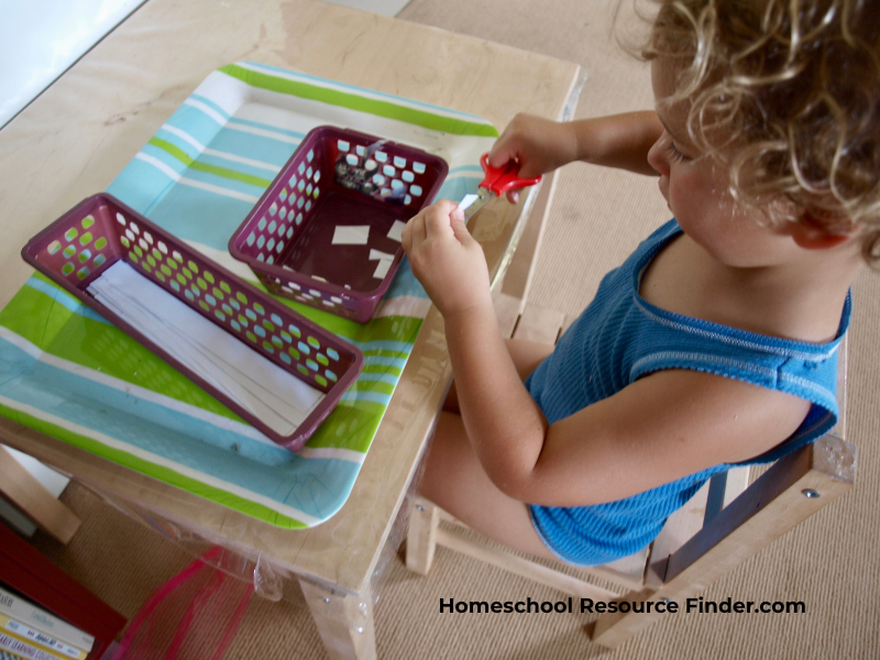 Homeschooling with a toddler. Image of a toddle practicing scissor skills in the homeschool